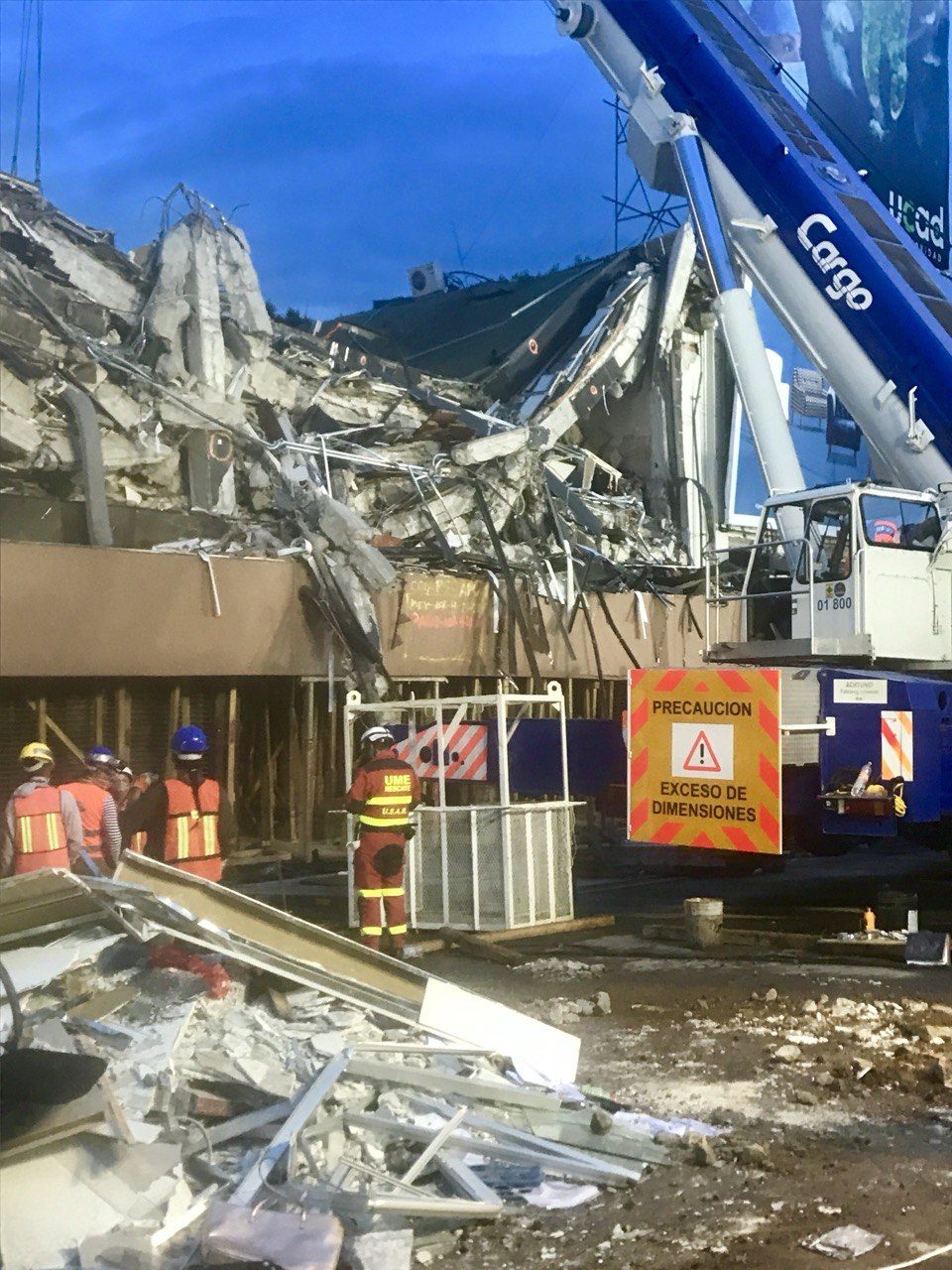 Pancake-collapsed building with a blue crane and rescuers in orange vests, Mexico City 2017.